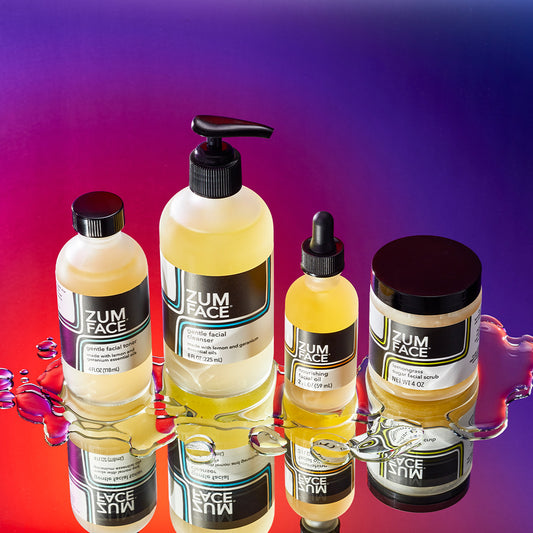 Four labeled Zum Face products lined up on a mirrored surface with water droplets surrounding them. Purple to orange thermal gradient background. Products shown left to right: bottle of Gentle Facial Toner, a Gentle Facial Cleanser bottle with a pump, a Nourishing Facial Oil bottle with a dropper, and Lemongrass Sugar Facial Scrub jar.