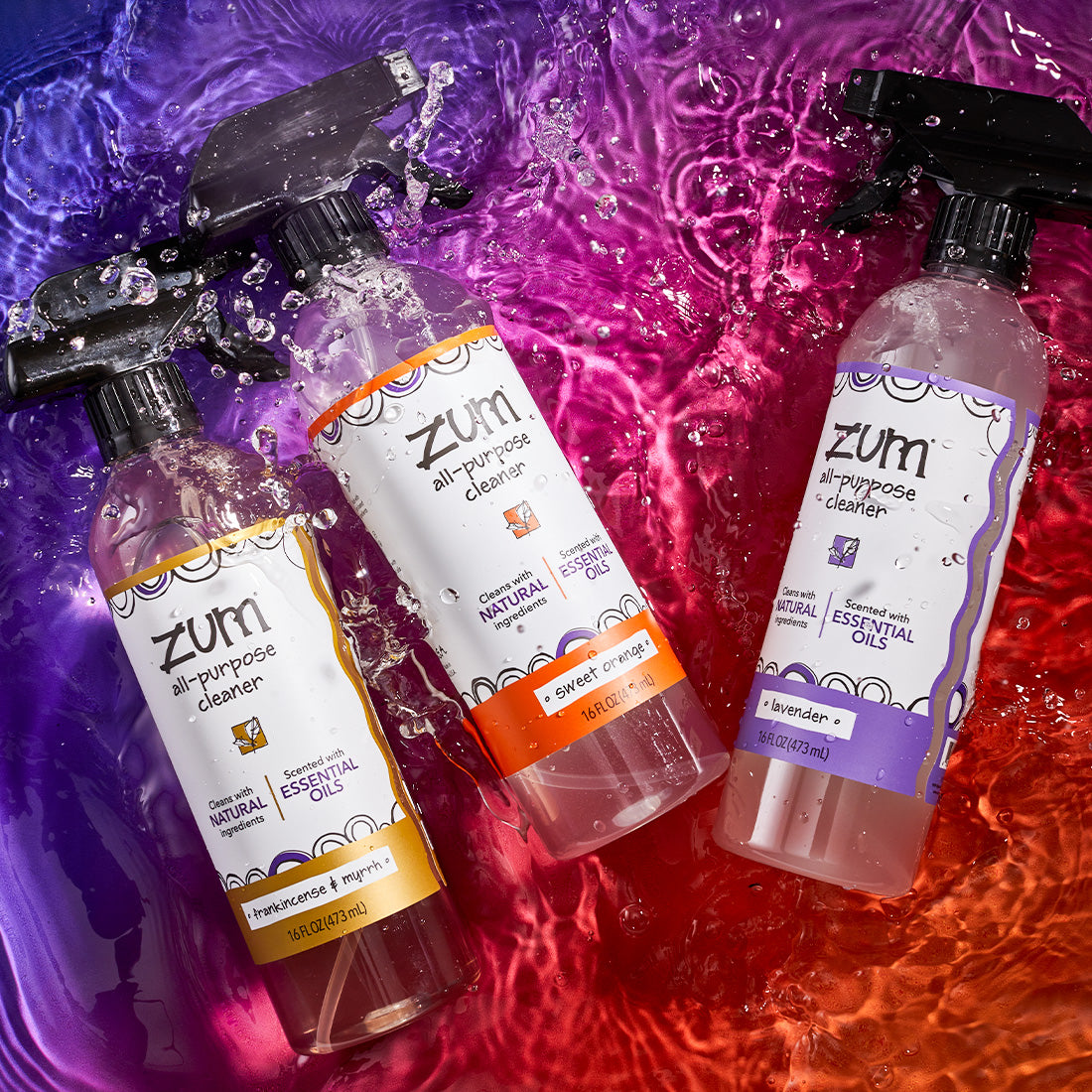 Labeled Frankincense & Myrrh, Sweet Orange, and Lavender all-purpose cleaner bottles with a sprayer. Splashing in a watered surface with a purple to orange gradient background.