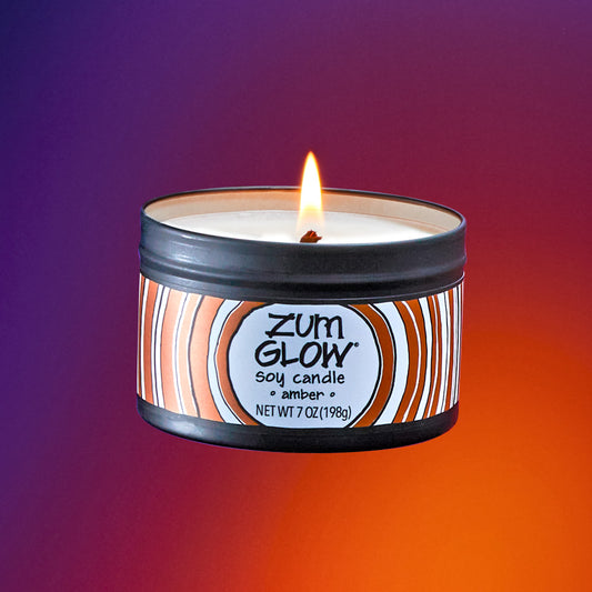 A lit labeled soy candle in a black tin flying in the air. Purple to orange gradient background.