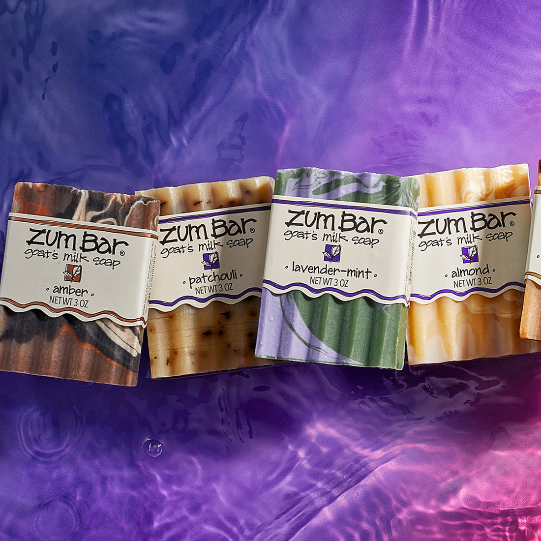 Amber, Patchouli, Lavender-Mint, and Almond labeled bar soaps lined up on a watered surface. Purple to pink gradient background.