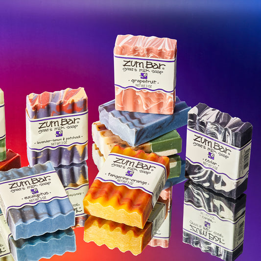 Various labeled bar soaps laying in angles and standing on unlabeled bar soaps on a mirrored surface. The scents present are Eucalyptus, Lavender-Lemon & Patchouli, Tangerine-Orange, Grapefruit, and Cedar. Purple to orange thermal gradient background.