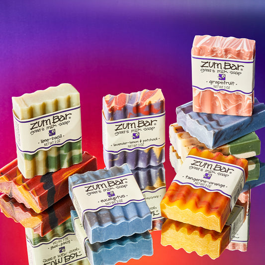 Various labeled bar soaps laying in angles and standing on unlabeled bar soaps on a mirrored surface. The scents present are Lime-Basil, Eucalyptus, Lavender-Lemon & Patchouli, Tangerine-Orange, and Grapefruit. Purple to orange thermal gradient background.