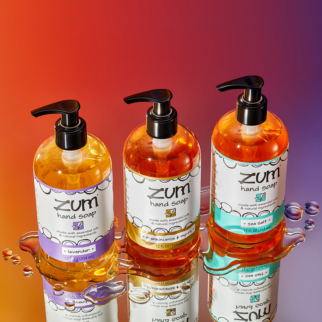 Three Hand Soap bottles with pumps lined up diagonally on a mirrored sruface with water droplets surrounding them. Scents are Lavender, Frankincense & Myrrh, and Sea Salt. Orange to purple gradient background.