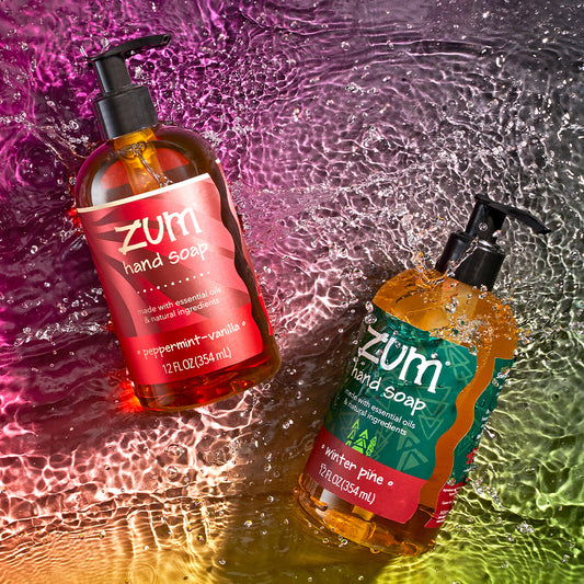 Peppermint-Vanilla and Winter Pine Hand Soap bottles with pumps splashing on a watered surface. Pinkish red to green gradient background.