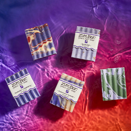 Lavender-Rosemary, Lavender-Lemon & Patchouli, Frankincense-Lavender, Lavender, and Lavender-Mint scented waxy bar soaps laying on a watered surface. Purple to orange gradient background