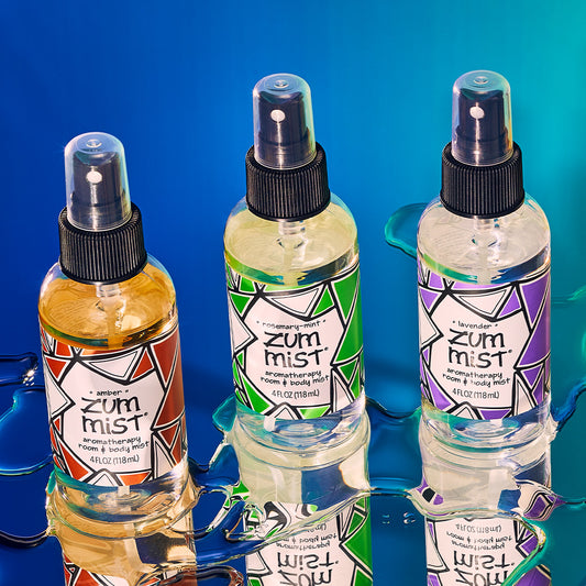 Amber, Rosemary-Mint, and Lavender room & body mist bottles on a mirrored surface diagonally. Water surrounds the bottom of the bottles. Blue gradient background.