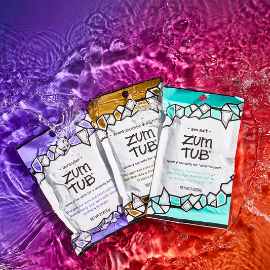 Lavender, frankincense & myrrh, and sea salt scented bath salts in packets splashing on a watered surface. purple to orange background.