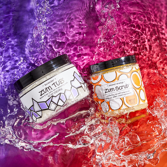 Jarred lavender epsom and sea salt bath salts and almond-orange sea salt body scrub in splashing water. Purple to orange background.