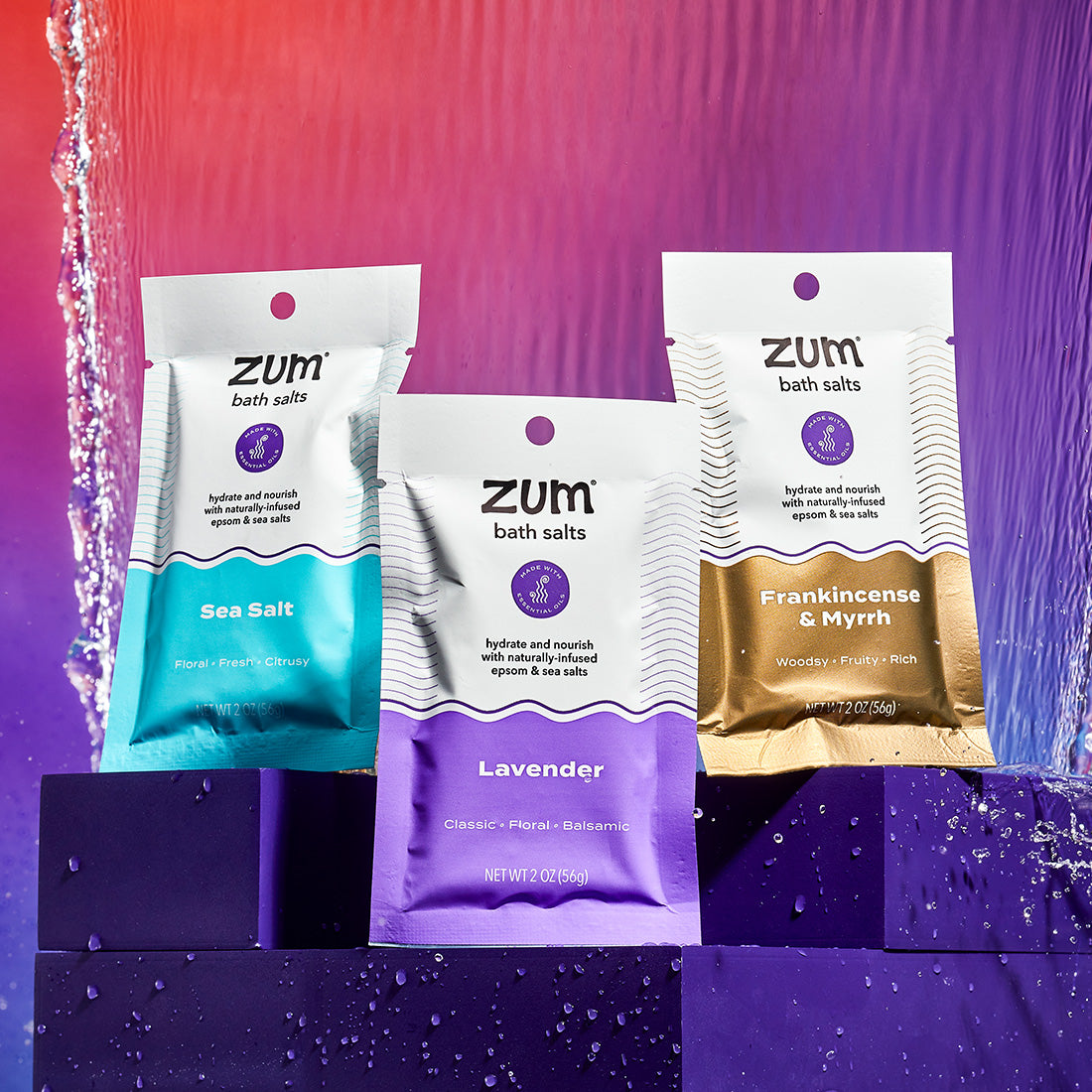 Sea Salt, Lavender, and Frankincense & Myrrh scented Bath Salt packets on purple risers with a waterfall behind. Orange to purple gradient background.