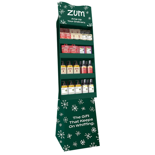 A freestanding green retail display stand with white snowflake graphics and the text 'ZUM Amp Up Your Ordinary.' on a white background. This shipper includes an assort of products in Peppermint-Vanilla, Holiday Almond, and Frosted Fir Scent.